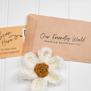 Op de afbeelding: Een wit en bruin gehaakte bloem voor een beige envelop met het opschrift Our Friendly World Crafting Sustainability en een lichtoranje kaart met de tekst Scan Here to view your crochet pattern en een QR-code.