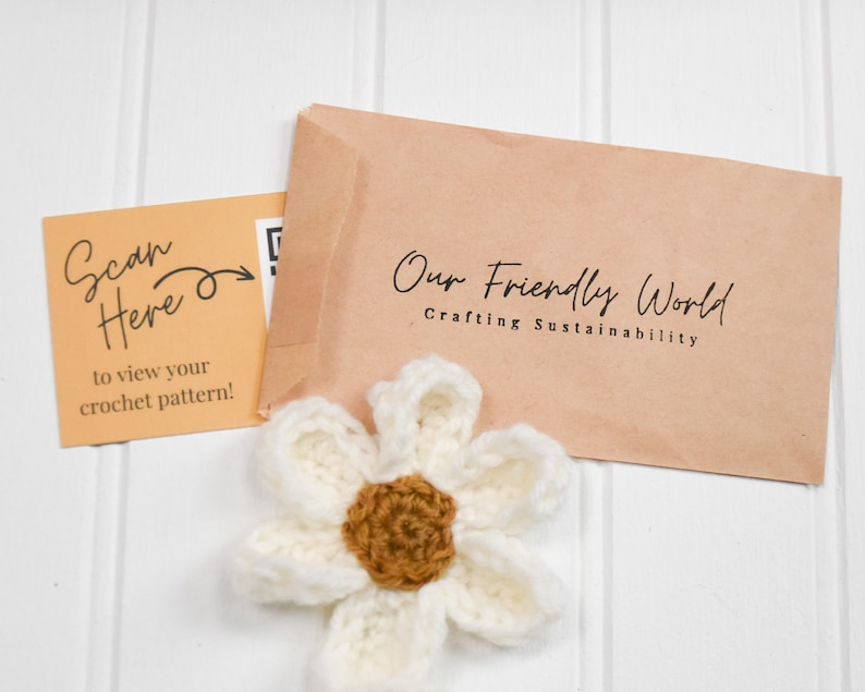 Op de afbeelding: Een wit en bruin gehaakte bloem voor een beige envelop met het opschrift Our Friendly World Crafting Sustainability en een lichtoranje kaart met de tekst Scan Here to view your crochet pattern en een QR-code.
