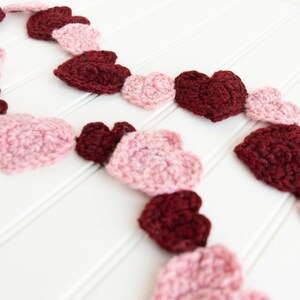 May include: A garland of crocheted hearts in pink and burgundy shades. The hearts are strung together to create a festive decoration.