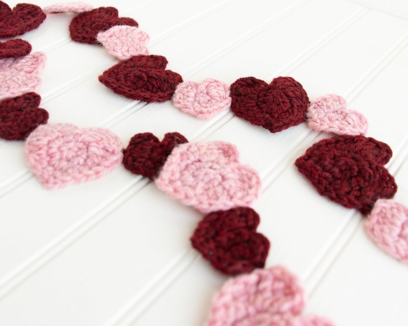 May include: A garland of crocheted hearts in pink and burgundy shades. The hearts are strung together to create a festive decoration.