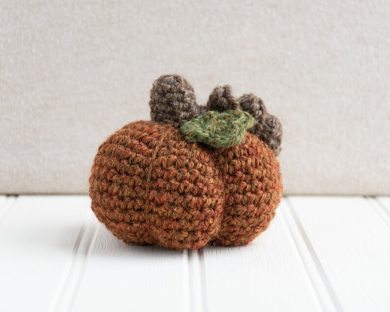May include: A crocheted pumpkin with a brown stem and green leaves. The pumpkin is made of a brown yarn and has a textured surface.
