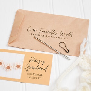 Op de afbeelding: Een knutselpakket met een bruine papieren envelop met het opschrift Our Friendly World Crafting Sustainability, een naainaald, een zwarte steekmarkeerder en een kaart met Daisy Garland Eco-Friendly Crochet Kit.