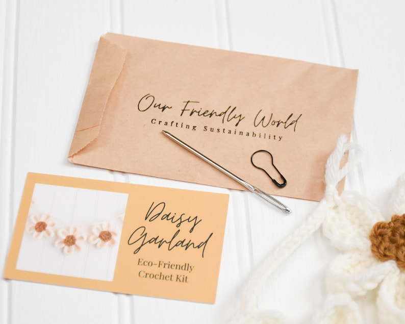 Op de afbeelding: Een knutselpakket met een bruine papieren envelop met het opschrift Our Friendly World Crafting Sustainability, een naainaald, een zwarte steekmarkeerder en een kaart met Daisy Garland Eco-Friendly Crochet Kit.