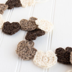 May include: A garland of crocheted hearts in brown, beige and white. The hearts are strung together and hang against a white background.