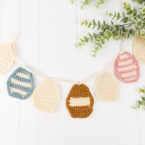 May include: A crocheted Easter egg garland against a white background. The garland is made of different coloured eggs, including cream, brown, pink, and blue. The eggs are decorated with white stripes.