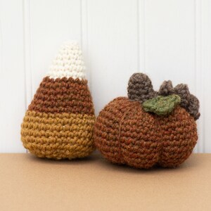 May include: A crocheted candy corn and a crocheted pumpkin. The candy corn is brown, yellow, and white. The pumpkin is brown with a green leaf.