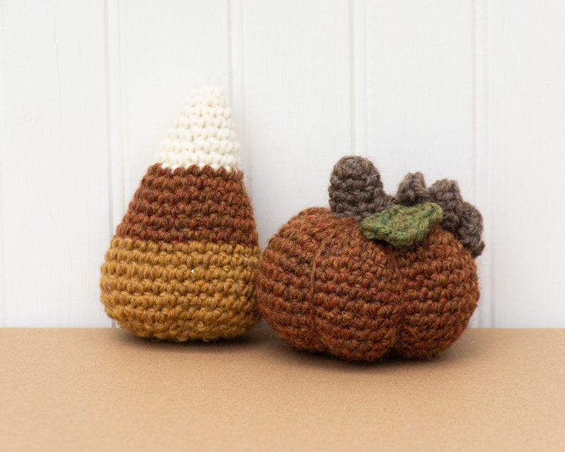 May include: A crocheted candy corn and a crocheted pumpkin. The candy corn is brown, yellow, and white. The pumpkin is brown with a green leaf.