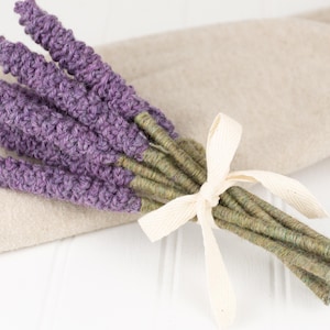 May include: A bundle of lavender stems with purple flower heads, tied with a cream-colored ribbon. The stems are green and the lavender is resting on a beige cloth.