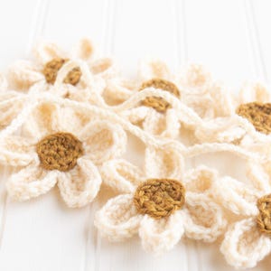 Op de afbeelding: Een gehaakte slinger met meerdere madeliefvormige bloemen verbonden door een garenkoord. Elke bloem heeft cr&egrave;mekleurige bloemblaadjes en een rond bruin hart. De afzonderlijke bloemen zijn met elkaar verbonden in een doorlopende streng.