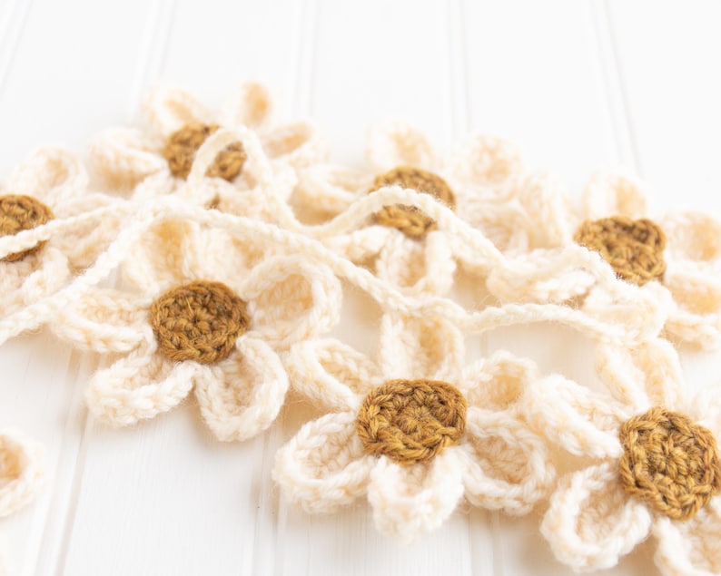 Op de afbeelding: Een gehaakte slinger met meerdere madeliefvormige bloemen verbonden door een garenkoord. Elke bloem heeft cr&egrave;mekleurige bloemblaadjes en een rond bruin hart. De afzonderlijke bloemen zijn met elkaar verbonden in een doorlopende streng.