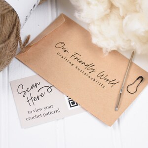 Puede incluir: Un sobre de papel marr&oacute;n con el texto "Our Friendly World Crafting Sustainability" impreso en &eacute;l. Una tarjeta blanca con el texto "Scan Here to view your crochet pattern!" y un c&oacute;digo QR est&aacute; al lado del sobre. Tambi&eacute;n hay una madeja de hilo marr&oacute;n, una aguja y un imperdible en la imagen.