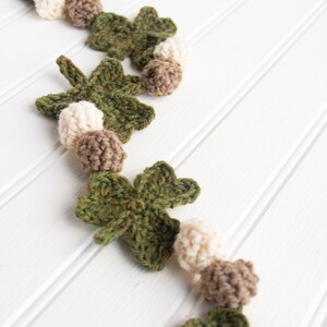 Puede incluir: Una guirnalda de crochet con hojas de tr&eacute;bol verdes y bolas marrones y blancas. La guirnalda est&aacute; colgada en una pared blanca.