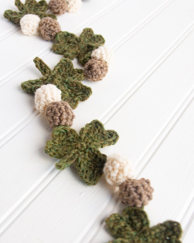 Puede incluir: Una guirnalda de crochet con hojas de tr&eacute;bol verdes y bolas marrones y blancas. La guirnalda est&aacute; colgada en una pared blanca.