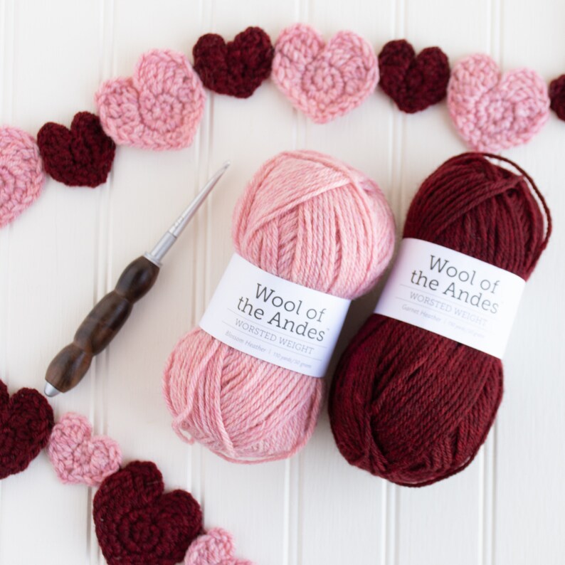 May include: Two balls of yarn, one pink and one burgundy, with the label "Wool of the Andes Worsted Weight". A crochet hook and a garland of pink and burgundy crocheted hearts are also pictured.