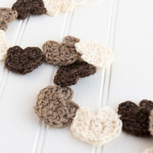 May include: A garland of crocheted hearts in brown, beige and white. The hearts are strung together and hang against a white background.