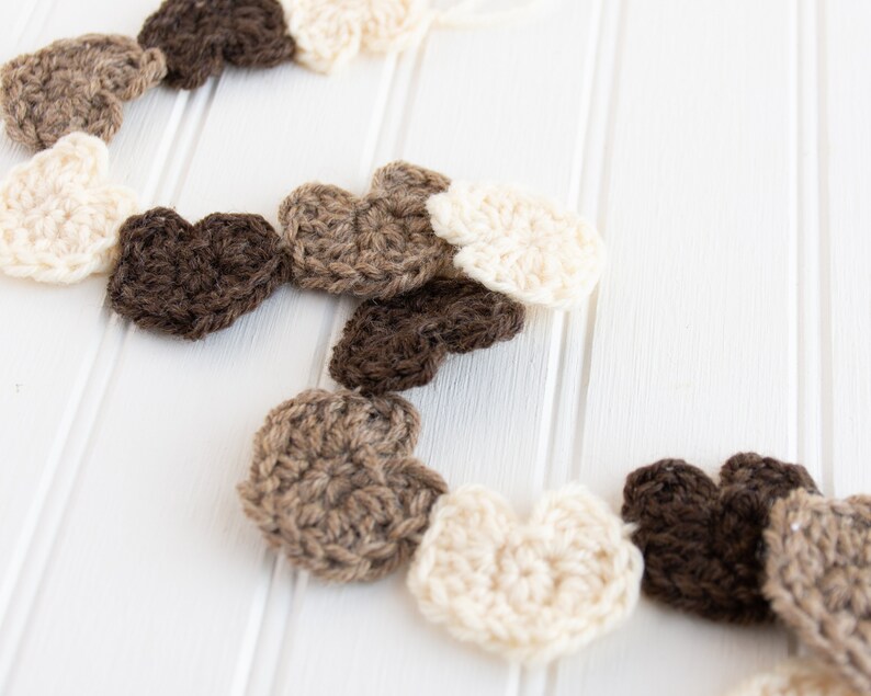 May include: A garland of crocheted hearts in brown, beige and white. The hearts are strung together and hang against a white background.