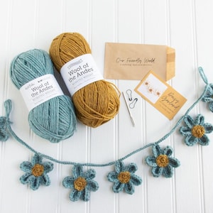 Op de afbeelding: Twee bollen Wool of the Andes worsted weight garen in blauwgroen en mosterdgeel, een gehaakte madeliefjesslinger, een metalen naald, twee steekmarkeerders, een bruine envelop met Our Friendly World en een kaart met Daisy Garland.