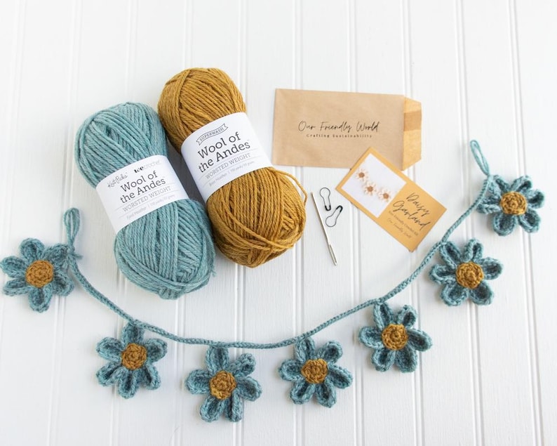 Op de afbeelding: Twee bollen Wool of the Andes worsted weight garen in blauwgroen en mosterdgeel, een gehaakte madeliefjesslinger, een metalen naald, twee steekmarkeerders, een bruine envelop met Our Friendly World en een kaart met Daisy Garland.