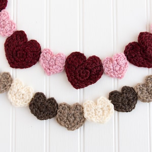 May include: A garland of crocheted hearts in various colours, including pink, red, brown, and cream, hanging on a white wooden background.