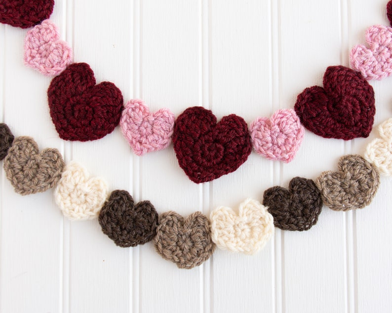 May include: A garland of crocheted hearts in various colours, including pink, red, brown, and cream, hanging on a white wooden background.