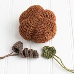 May include: A crocheted pumpkin with brown, green, and orange yarn. The pumpkin is made up of five lobes and has a stem made of brown yarn. There are two separate crocheted leaves made of green yarn.