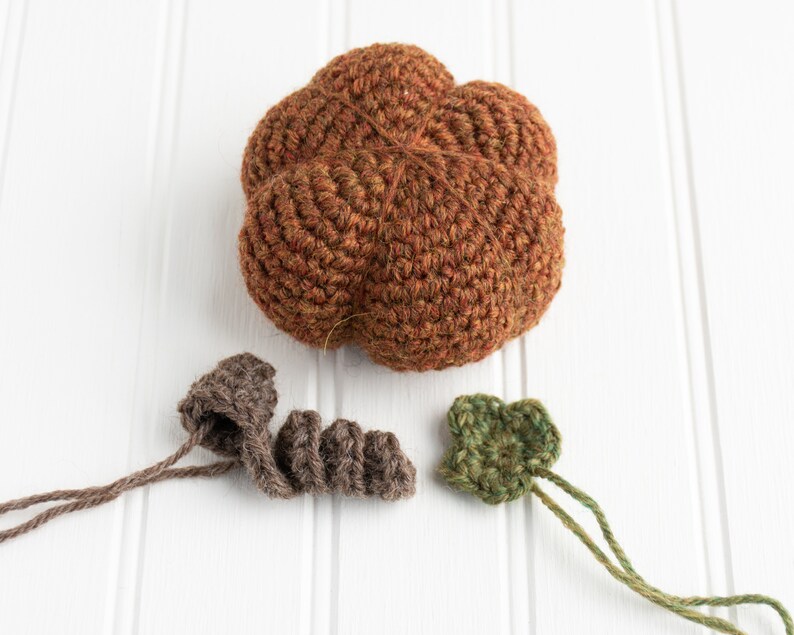 May include: A crocheted pumpkin with brown, green, and orange yarn. The pumpkin is made up of five lobes and has a stem made of brown yarn. There are two separate crocheted leaves made of green yarn.