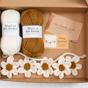 Op de afbeelding: Een haakpakket in een kartonnen doos met een bol witte en een bol bruine wol, een metalen naald, een steekmarkeerder en een kaart met de tekst Daisy Garland Eco-Friendly Crochet Kit en Our Friendly World.