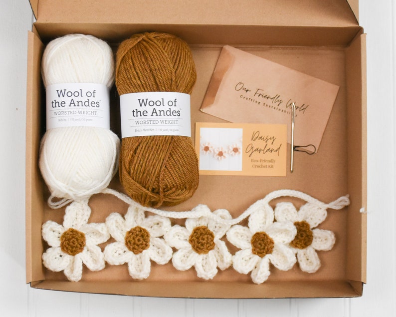 Op de afbeelding: Een haakpakket in een kartonnen doos met een bol witte en een bol bruine wol, een metalen naald, een steekmarkeerder en een kaart met de tekst Daisy Garland Eco-Friendly Crochet Kit en Our Friendly World.