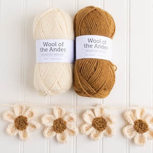 Op de afbeelding: Twee bollen Wool of the Andes garen in cr&egrave;me en bruin, geplaatst boven een gehaakte slinger met cr&egrave;me-kleurige bloemblaadjes en bruine harten. Elk label op het garen bevat de tekst Wool of the Andes.