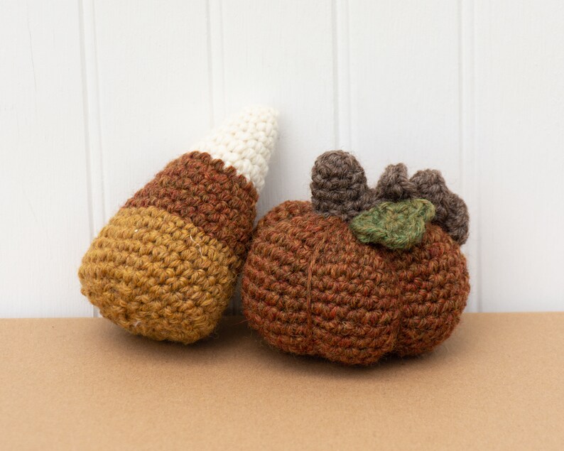 May include: Two crocheted fall decorations: a brown and white candy corn and a brown pumpkin with a green leaf.