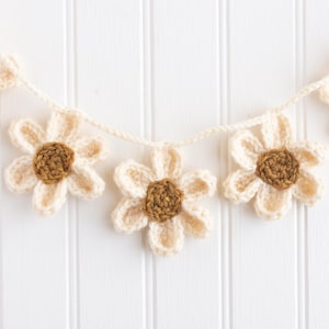 Op de afbeelding: Een gehaakte slinger met vijf individuele bloemen verbonden door een cr&egrave;mekleurig garenkoord. Elke bloem heeft zes cr&egrave;mekleurige bloemblaadjes rondom een bruin, cirkelvormig centrum, met een duidelijk zichtbare garenstructuur.