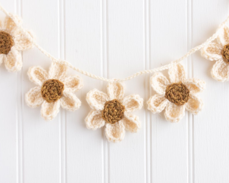 Op de afbeelding: Een gehaakte slinger met vijf individuele bloemen verbonden door een cr&egrave;mekleurig garenkoord. Elke bloem heeft zes cr&egrave;mekleurige bloemblaadjes rondom een bruin, cirkelvormig centrum, met een duidelijk zichtbare garenstructuur.