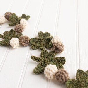 Puede incluir: Una guirnalda de crochet con hojas verdes y bolas blancas y marrones. La guirnalda est&aacute; sobre un fondo de madera blanca.