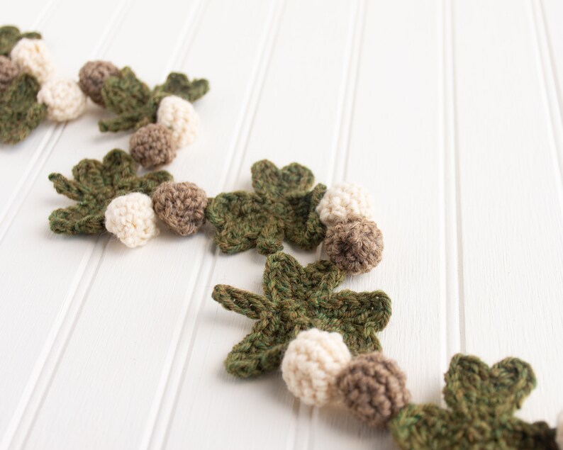 Puede incluir: Una guirnalda de crochet con hojas verdes y bolas blancas y marrones. La guirnalda est&aacute; sobre un fondo de madera blanca.