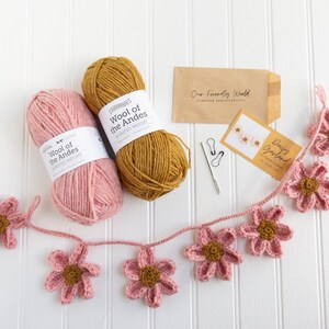 Op de afbeelding: Twee bollen Wool of the Andes garen van gemiddeld gewicht in roze en mosterdgeel, een gehaakte madeliefjesslinger, twee steekmarkeerders, een metalen naald en twee kleine papieren enveloppen met het label Our Friendly World en Daisy Garland.
