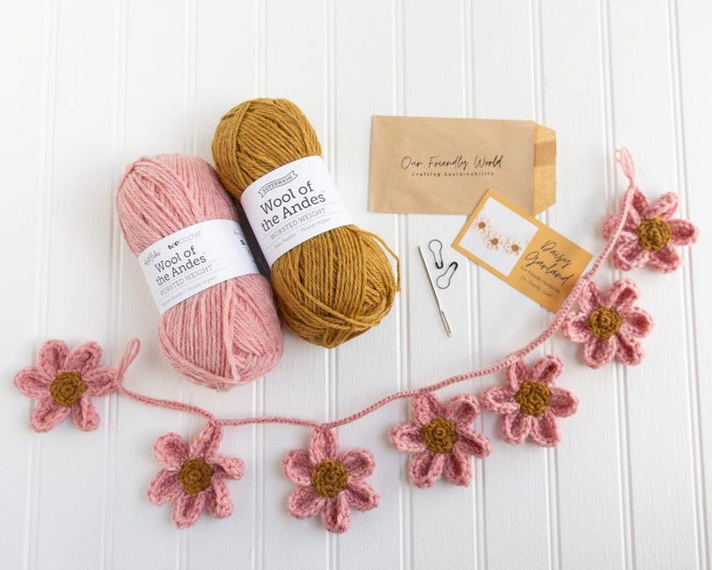 Op de afbeelding: Twee bollen Wool of the Andes garen van gemiddeld gewicht in roze en mosterdgeel, een gehaakte madeliefjesslinger, twee steekmarkeerders, een metalen naald en twee kleine papieren enveloppen met het label Our Friendly World en Daisy Garland.