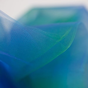 Könnte beinhalten: Nahaufnahme eines blauen und grünen, durchsichtigen Stoffes mit einer zarten, gewebten Textur. Der Stoff ist drapiert und gefaltet und erzeugt weiche, fließende Linien.