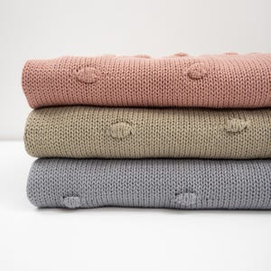 Könnte beinhalten: Drei gestrickte Decken übereinander gestapelt. Die oberste Decke ist hellrosa, die mittlere Decke ist hellbraun und die unterste Decke ist hellgrau. Alle drei Decken haben ein strukturiertes Muster mit kleinen erhabenen Kreisen.