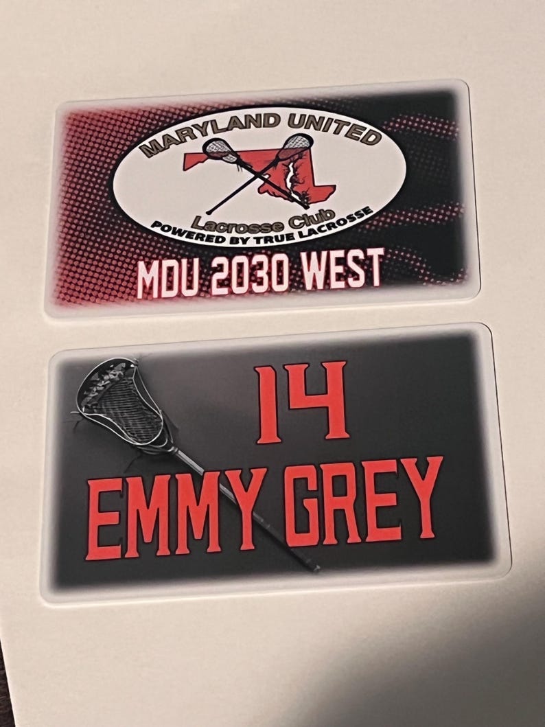Może przedstawiać: Dwie prostokątne naklejki o tematyce lacrosse. G&oacute;rna naklejka zawiera tekst "MARYLAND UNITED" i "Lacrosse Club" z grafiką kija do lacrosse. Dolna naklejka wyświetla "14" i "EMMY GREY" z kijem do lacrosse.