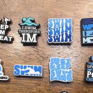 May include: Ten colorful shoe charms with swimming-themed designs. The charms feature phrases like "Eat Sleep Swim Repeat", "I Swim Therefore IM", "Just Keep Swimming", and "Life is Cool in the Pool".