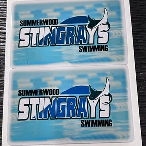 Może przedstawiać: Dwie prostokątne naklejki z jasnoniebieskim tłem. Zawierają tekst "SUMMERWOOD STINGRAYS SWIMMING" i stylizowany wz&oacute;r płaszczki. Naklejki są na białym tle.