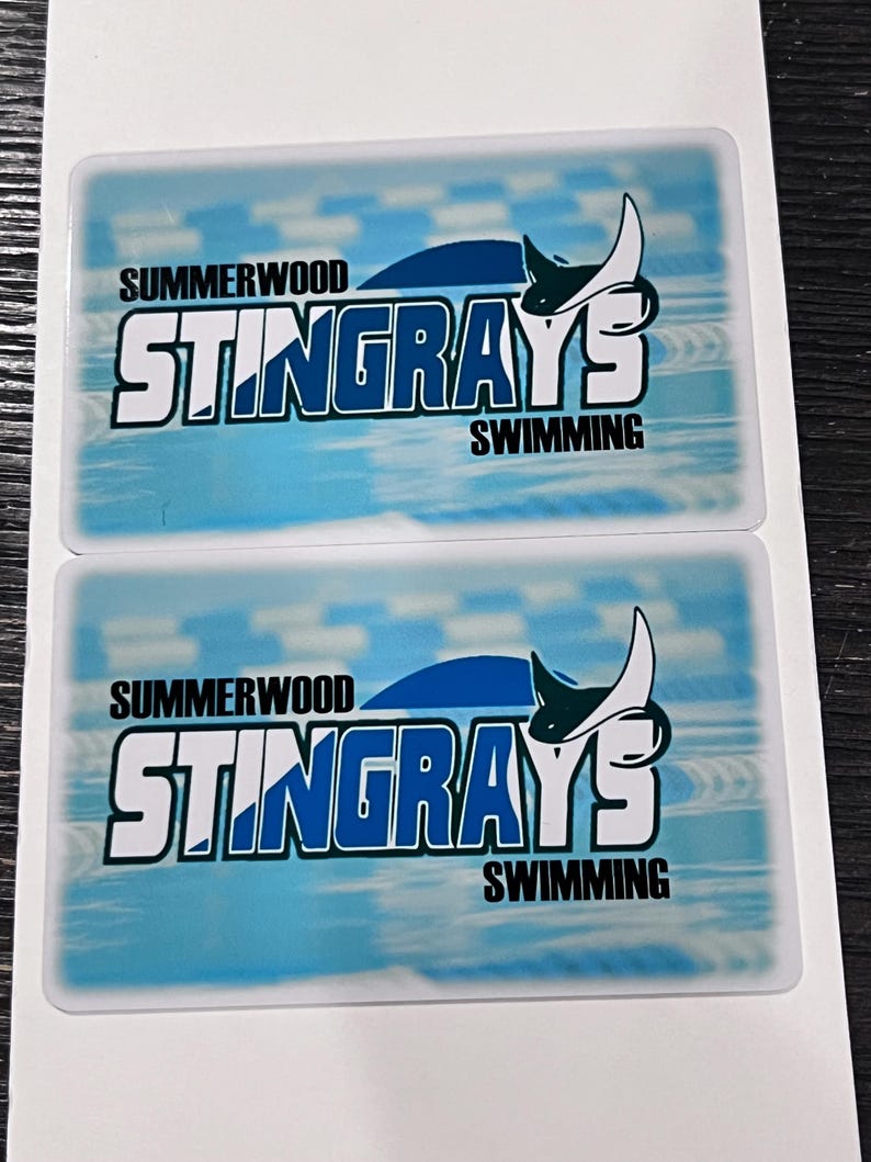 Może przedstawiać: Dwie prostokątne naklejki z jasnoniebieskim tłem. Zawierają tekst "SUMMERWOOD STINGRAYS SWIMMING" i stylizowany wz&oacute;r płaszczki. Naklejki są na białym tle.