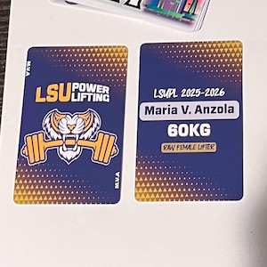 Może przedstawiać: Dwie prostokątne karty z granatowym tłem i złotymi akcentami. Jedna karta zawiera tekst "LSU Power Lifting" z tygrysem i grafiką sztangi. Druga karta wyświetla "LSUPL 2025-2026 Maria V. Anzola 60KG Raw Female Lifter."