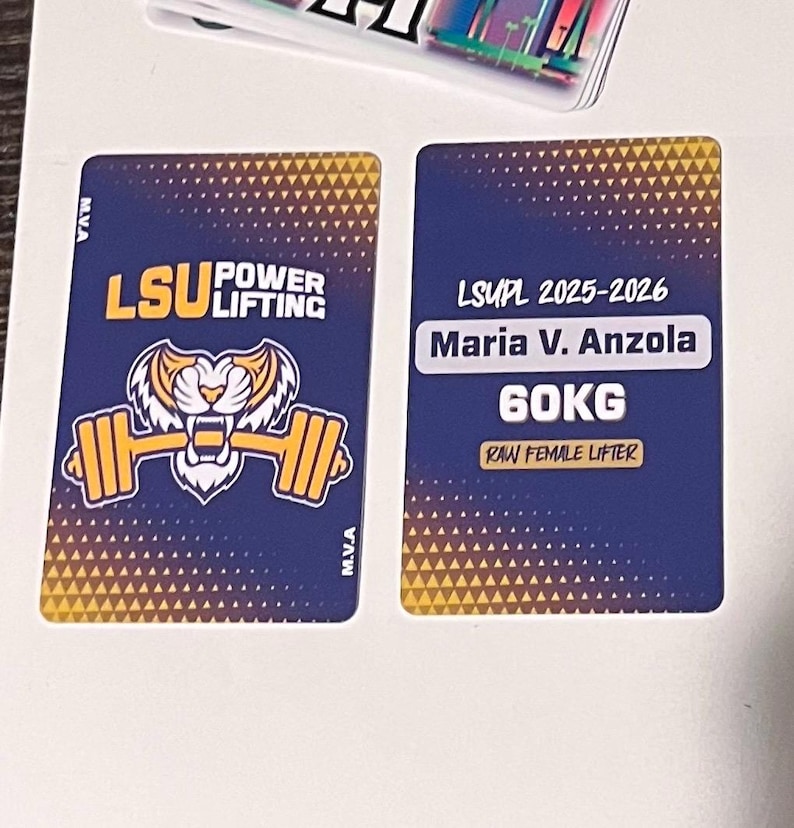 Może przedstawiać: Dwie prostokątne karty z granatowym tłem i złotymi akcentami. Jedna karta zawiera tekst "LSU Power Lifting" z tygrysem i grafiką sztangi. Druga karta wyświetla "LSUPL 2025-2026 Maria V. Anzola 60KG Raw Female Lifter."