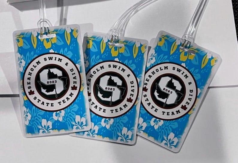 Może przedstawiać: Trzy prostokątne etykiety bagażowe z niebieskim tłem w kwiatowy wz&oacute;r. Każda etykieta ma okrągły emblemat z napisem "SEAHOLM SWIM & DIVE STATE TEAM 2023". Etykiety mają przezroczyste plastikowe pętle.