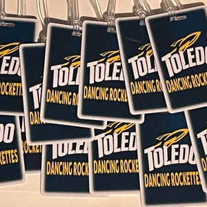 Może przedstawiać: Kolekcja prostokątnych etykiet bagażowych z napisem "TOLEDO DANCING ROCKETTES" w białych i złotych literach. Etykiety mają granatowe tło i grafikę rakiety. Każda etykieta ma przezroczystą plastikową pętlę.
