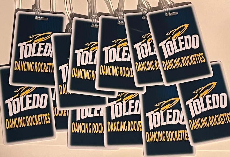 Może przedstawiać: Kolekcja prostokątnych etykiet bagażowych z napisem "TOLEDO DANCING ROCKETTES" w białych i złotych literach. Etykiety mają granatowe tło i grafikę rakiety. Każda etykieta ma przezroczystą plastikową pętlę.