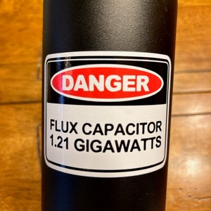 May include: Black water bottle with a white warning label that reads "DANGER" in a red circle and "FLUX CAPACITOR 1.21 GIGAWATTS" in black text.