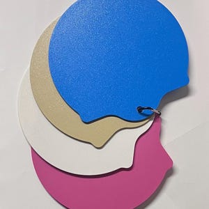 May include: A set of four circular, speech-bubble-shaped swatches in blue, beige, white, and magenta. Each swatch has a textured surface and a small tab on the side. The swatches are stacked and held together by a small black metal ring.