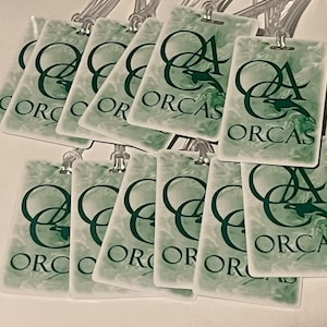 Może przedstawiać: Kolekcja prostokątnych etykiet bagażowych z zielono-białym marmurkowym wzorem. Każda etykieta zawiera napis "ORCAS" oraz grafikę orki w ciemnozielonym kolorze. Etykiety posiadają przezroczyste plastikowe pętle do mocowania.